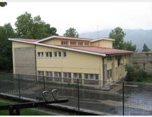 Edificio a due piani color giallo con tetto rosso a falde e file di finestre, circondato da una recinzione metallica e da un cortile asfaltato. Sullo sfondo si vedono alberi e colline o montagne parzialmente coperte da foschia.
