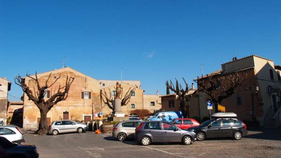 Piazza urbana lastricata con parcheggio di auto, circondata da edifici in muratura dai toni ocra e da alberi potati senza foglie. Al centro si vede una piccola aiuola con un monumento, sotto un cielo sereno.