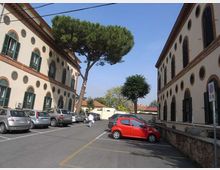 Cortile-parcheggio tra due edifici a più piani con facciate chiare, finestre ad arco e piccoli oblò circolari lungo le pareti. Al centro si vedono alberi alti e diverse auto parcheggiate, con strada asfaltata e segnaletica a terra.