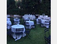 Spazio all’aperto su prato con tavoli rotondi coperti da tovaglie bianche e sedie pieghevoli nere, disposto per un evento. Sullo sfondo si vedono alberi e siepi che delimitano l’area.