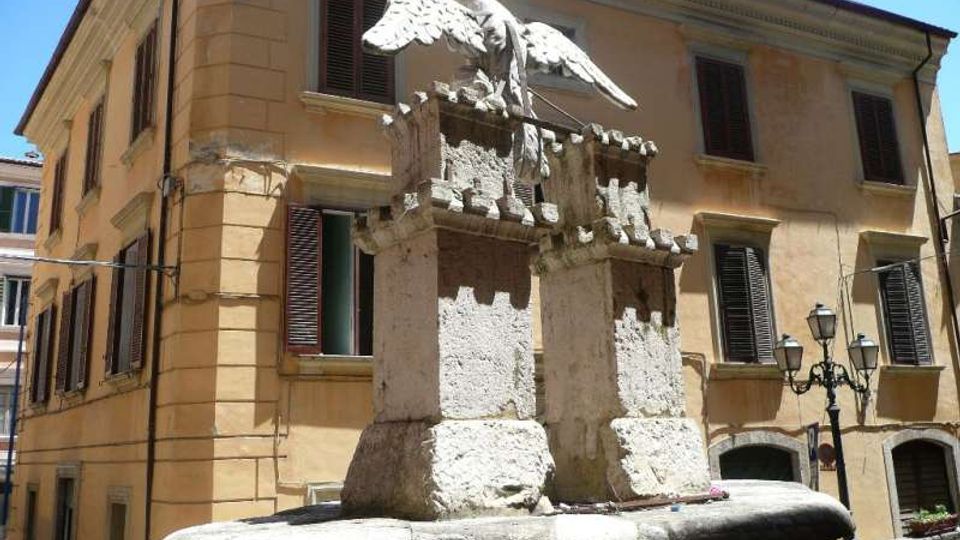 Fontana in pietra al centro di una piccola piazza, con basamento circolare e due pilastri sormontati da una scultura di aquila con le ali spiegate. Sullo sfondo si vede un edificio storico color ocra con persiane scure e lampioni in ferro.