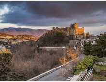 Castello in pietra su un’altura, con torri e mura illuminate, che domina una valle boscosa. In primo piano una strada curva con muretto e alcune case ai margini del pendio, sullo sfondo montagne sotto un cielo nuvoloso.
