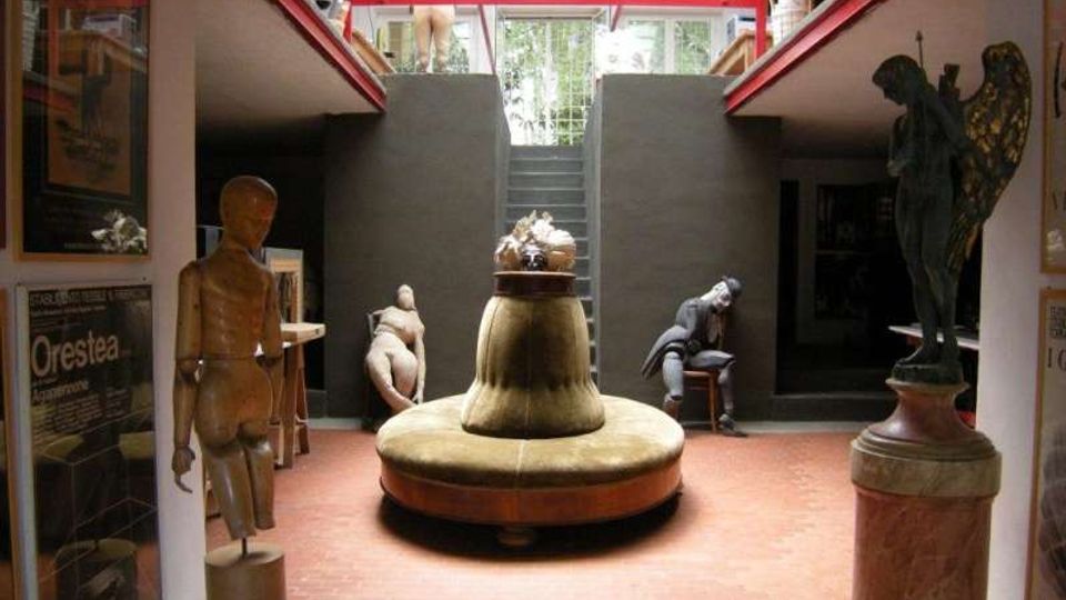 Interno di un museo o spazio espositivo con pavimento in mattoni rossi, una grande struttura circolare centrale e statue esposte su piedistalli ai lati. Sul fondo si vede una scalinata che sale verso un’apertura superiore con ringhiera e luce naturale.