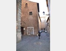 Vicolo urbano lastricato con un grande edificio in mattoni a vista sulla sinistra, con finestre piccole e una porta murata. La strada stretta prosegue tra palazzi storici con un lampione sospeso e alcune auto parcheggiate sullo sfondo.