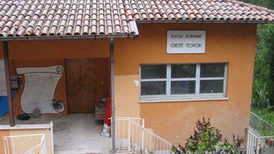 Edificio scolastico a un piano con pareti color ocra e tetto in tegole, con ingresso coperto e rampa con ringhiera bianca. Sulla facciata si vedono una fila di finestre e un’insegna con la scritta “Scuola elementare Oreste Teodori”, immerso in un contesto verde.