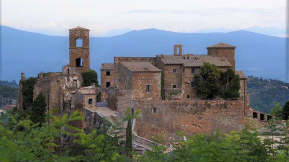 Complesso fortificato in pietra su un crinale collinare, con mura e edifici in muratura e una torre campanaria quadrata con aperture ad arco. Sullo sfondo si vedono montagne azzurrate e cielo chiaro, con vegetazione in primo piano.