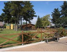 Parco urbano con ampio prato e alberi alti, delimitato da una staccionata in legno lungo un vialetto pavimentato con panchina. Sullo sfondo si vedono una piccola struttura in legno e un’area recintata con edifici vicini.