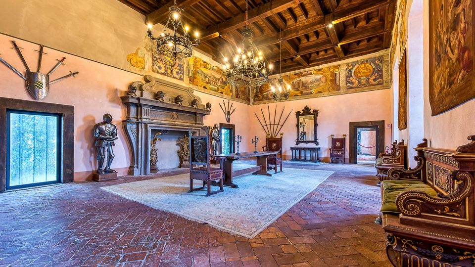 Ampia sala storica in stile rinascimentale con soffitto a travi di legno, grandi lampadari e pavimento in mattoni. Lungo le pareti ci sono un grande camino in pietra, armature decorative, affreschi e arredi d’epoca come sedie intagliate, tavoli e specchi.