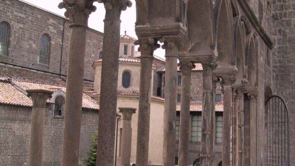 Loggiato in pietra di un edificio storico, con una fila di colonne sottili e archi decorati che affacciano su un cortile interno. Sullo sfondo si vedono facciate in muratura e tetti in tegole di altri edifici del complesso.
