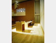 Interno di un’aula magna o sala conferenze con palco in legno, podio con microfono e lungo tavolo con sedie e postazioni. Pareti rivestite in pannelli di legno, balconata laterale e grandi porte sul fondo.