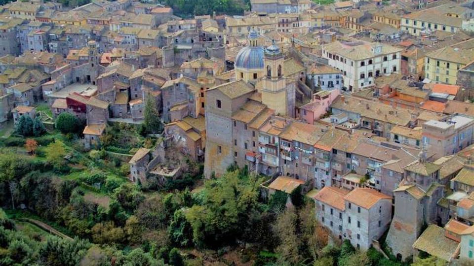 Veduta aerea di un borgo storico su pendio con case in pietra addossate e tetti in coppi, attraversato da strade strette. Al centro spicca una chiesa con cupola e campanile, circondata da vegetazione e terreni verdi ai margini dell’abitato.