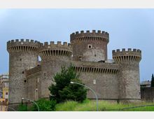 Castello medievale in pietra con quattro torri cilindriche merlate e bastioni, visto dall’esterno. In primo piano ci sono un’area verde con alberi e lampioni, con edifici urbani sullo sfondo sotto un cielo nuvoloso.
