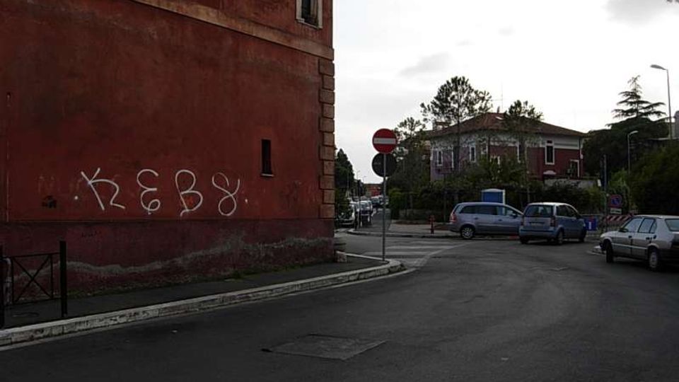 Incrocio urbano con ampia carreggiata asfaltata e alcune auto parcheggiate o in sosta, accanto a un grande edificio intonacato rosso con finestre ad arco e scritte di graffiti sul muro. Sullo sfondo si vedono segnaletica stradale, alberi e una casa a due piani con tetto a falde.