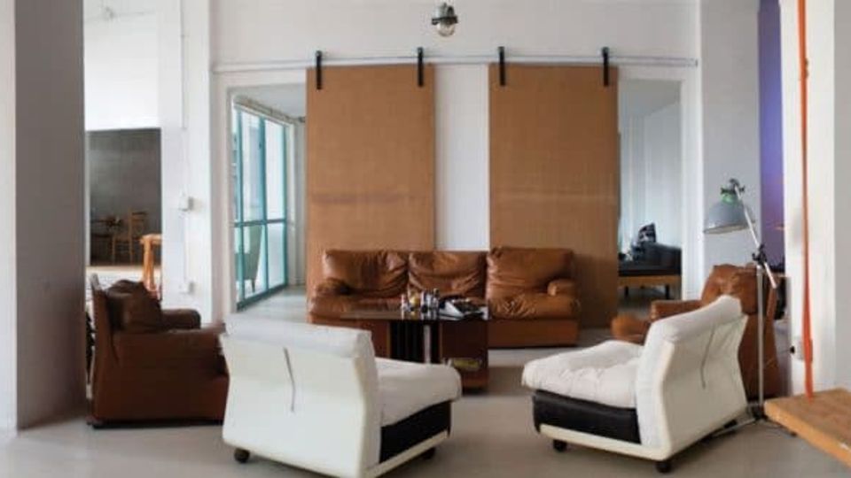 Ampio loft adibito a studio con grandi finestre industriali, pareti bianche e colonne, che include una zona ufficio con scrivanie e computer e un’area lounge con divani e poltrone. Sullo sfondo si vedono attrezzature e arredi da studio in un ambiente aperto e luminoso.