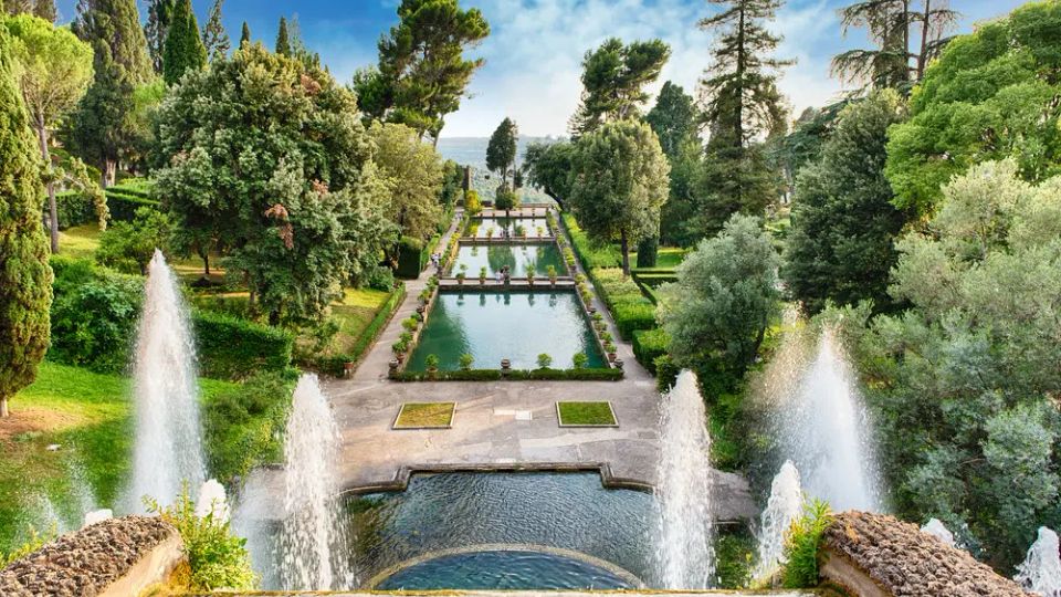 Veduta dall’alto di un giardino formale con vasche rettangolari in asse e percorsi simmetrici, circondato da alberi alti e siepi. In primo piano si vedono fontane a getto e una grande vasca, con il complesso che si estende verso l’orizzonte.