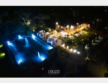 Veduta notturna dall’alto di una villa con giardino illuminato, area pavimentata e gazebo bianchi. In primo piano c’è una grande piscina rettangolare con fontane e luci blu, circondata da alberi e vialetti.