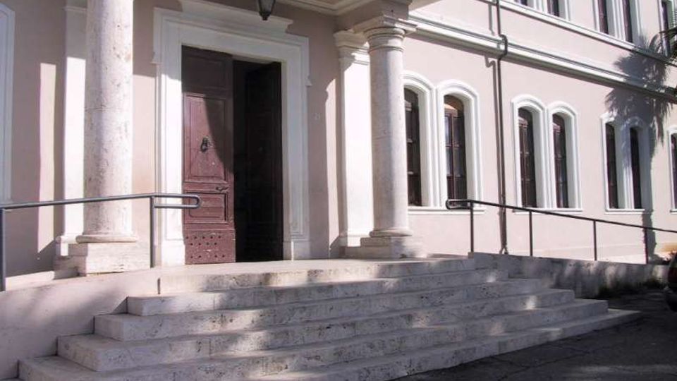 Ingresso di un edificio istituzionale con ampia scalinata in pietra e due colonne che sorreggono un portico sopra la porta principale. La facciata chiara presenta finestre ad arco e ringhiere metalliche lungo una rampa laterale.