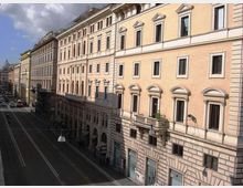 Veduta in prospettiva di una lunga strada urbana fiancheggiata da alti palazzi storici dai toni chiari, con facciate ornate da cornici e finestre regolari. La carreggiata è ampia e quasi vuota, con lampioni sospesi e cielo parzialmente nuvoloso sullo sfondo.