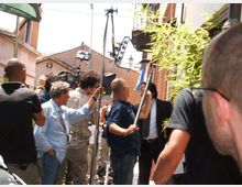 Ripresa in una stretta via urbana con edifici bassi e facciate in intonaco, attrezzature da set cinematografico come telecamera su supporto, aste e pannelli di illuminazione. Sul lato destro si vede una fitta pianta di bambù davanti a un edificio.