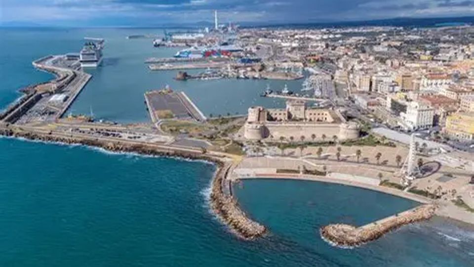 Veduta aerea di una città costiera con porto e moli, con navi ormeggiate e infrastrutture industriali sullo sfondo. In primo piano si vede un promontorio con un grande complesso fortificato in pietra e strade costiere che delimitano baie e frangiflutti sul mare.