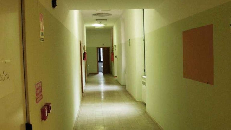 Corridoio interno lungo e stretto con pavimento piastrellato e pareti bicolore verde chiaro e bianco, illuminato da plafoniere a soffitto. Lungo le pareti si vedono porte e dispositivi antincendio, con una porta aperta in fondo al corridoio.