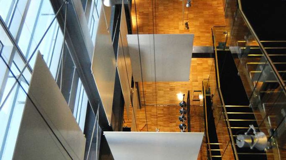 Interno di un atrio moderno a più piani visto dall’alto, con grandi vetrate verticali, pannelli bianchi sospesi e lampade a soffitto. Pavimento in parquet e passerelle con ringhiere in vetro e metallo lungo i lati.