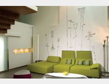 Soggiorno moderno con due divani chaise longue verdi e tavolino basso bianco su pavimento grigio. Parete bianca con decorazioni grafiche stilizzate, scala in legno a vista e nicchia illuminata con oggetti colorati.