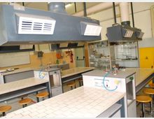 Laboratorio di chimica con banchi da lavoro rivestiti di piastrelle bianche, sgabelli e postazioni con rubinetti e attacchi per gas. Dal soffitto pendono grandi cappe aspiranti grigie, con armadi vetrati e scaffali sullo sfondo.