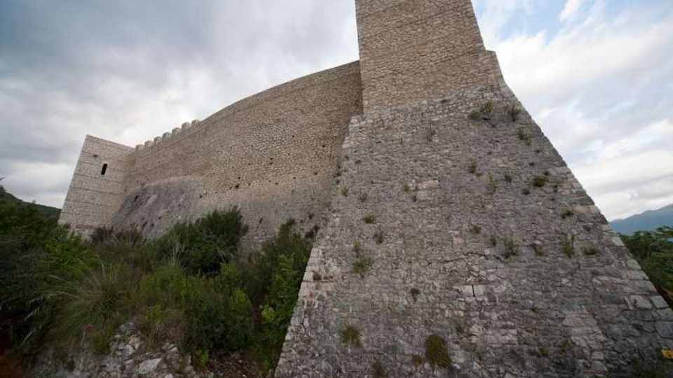 Mura esterne di un castello o fortezza in pietra con torre e bastioni inclinati, viste dal basso lungo un pendio roccioso. Vegetazione bassa intorno alla base e cielo nuvoloso sullo sfondo.