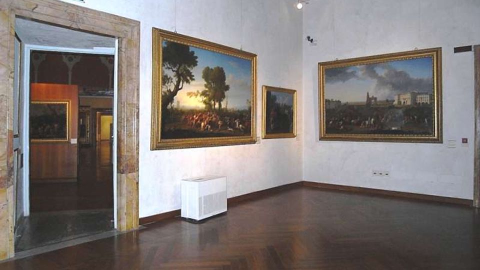 Sala di museo con pavimento in parquet scuro e pareti bianche, con grandi dipinti incorniciati appesi alle pareti e illuminazione a soffitto. A sinistra si vede un’apertura con cornice in pietra che conduce a un’altra galleria.