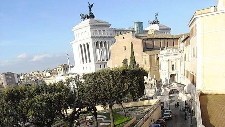 Veduta urbana con un grande monumento in marmo bianco a colonne, sormontato da statue equestri e una quadriga, sullo sfondo. In primo piano si vede una strada in discesa con auto parcheggiate, marciapiedi e alberi lungo un’area verde, affiancata da edifici storici.