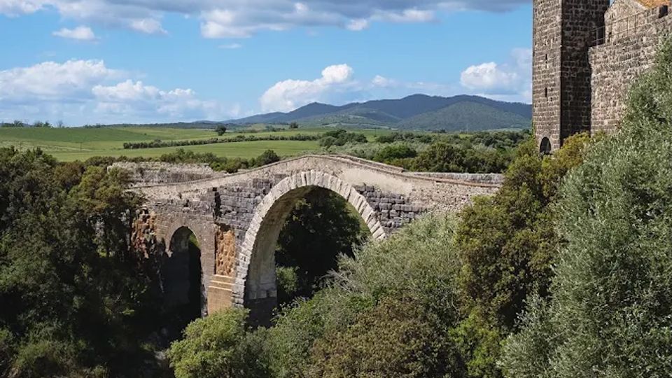 Ponte in pietra ad arco su una gola boscosa, con un fiume sul fondo. A destra si vede una torre in muratura tipo fortificazione, con colline e campi sullo sfondo sotto un cielo azzurro con grandi nuvole.