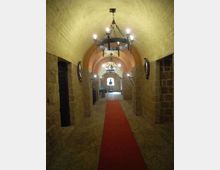 Corridoio sotterraneo in pietra con soffitto a volta e pavimento in mattoni, illuminato da grandi lampadari in ferro battuto. Un tappeto rosso corre lungo il centro fino a un arco in fondo, con nicchie e aperture laterali nelle pareti.