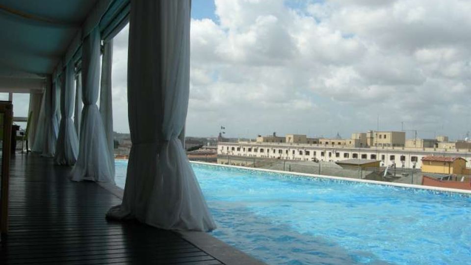 Terrazza con piscina a sfioro affiancata da una passerella in legno coperta, con tende bianche sospese lungo il lato. Sullo sfondo si vede lo skyline di edifici bassi della città sotto un cielo parzialmente nuvoloso.
