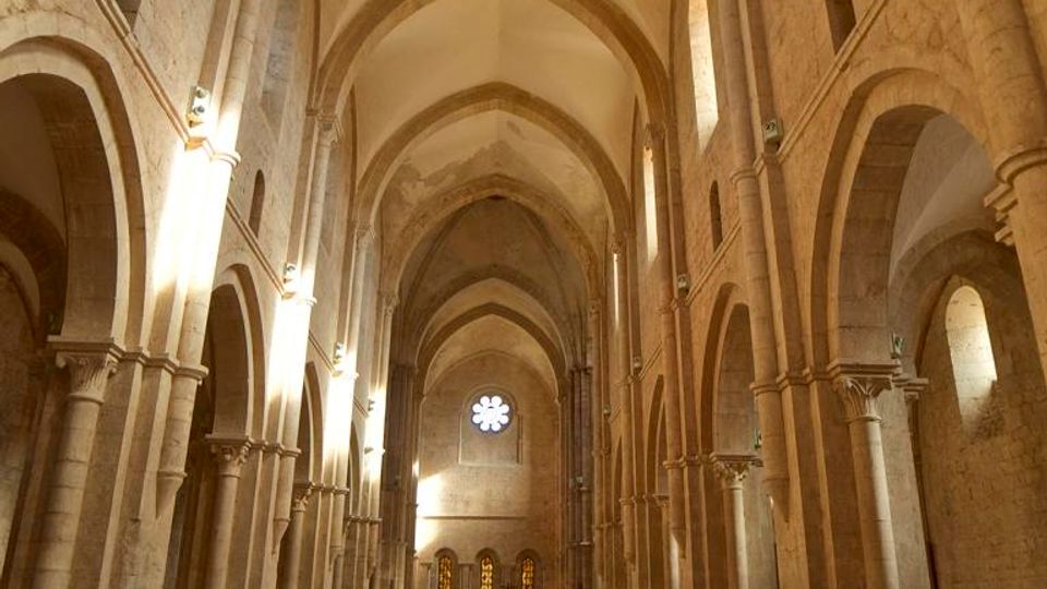 Interno di una chiesa in pietra con lunga navata centrale, arcate su colonne e soffitto a volte ogivali. File di panche in legno conducono verso l’abside, illuminata da una finestra a rosone e da strette finestre laterali.