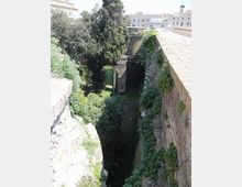 Veduta dall’alto di un fossato lungo mura in pietra e mattoni, con pareti parzialmente coperte di edera. In basso si vede vegetazione fitta con alberi e un piccolo passaggio ad arco sotto una struttura muraria.