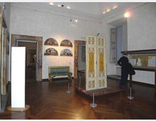 Sala museale con pavimento in parquet e pareti chiare, con un mobile decorato bianco e oro esposto su pedana delimitata da cordoni. Alle pareti si vedono dipinti ad arco, un divanetto e una vetrina con piccole sculture.