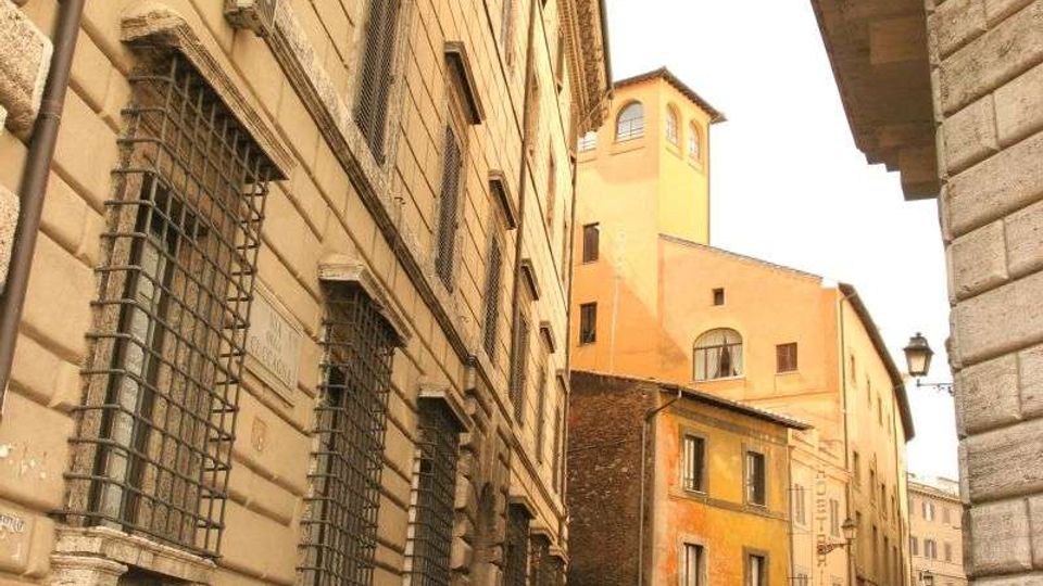 Strada stretta acciottolata in un centro storico, fiancheggiata da alti palazzi in pietra con finestre con inferriate e cornici modanate. Sullo sfondo si vedono edifici color ocra e un lampione, con una piccola piazzetta con siepi.