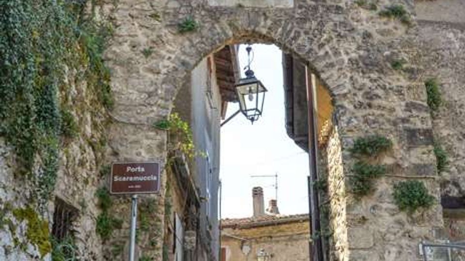 Antica porta urbana in pietra con arco a tutto sesto e merlature superiori, con targa e lanterna sospesa al centro dell’apertura. Sotto l’arco passa una stretta strada acciottolata tra edifici in muratura e pareti con vegetazione rampicante.