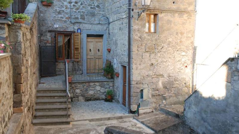 Cortile in un borgo con case in pietra grezza, piccola scalinata e portoncino con balconcino in ferro. Sul muro sono visibili una lanterna, finestre con persiane e alcuni vasi di fiori.