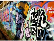 Muro esterno lungo una strada ricoperto da graffiti multicolori e street art, con grandi scritte e un personaggio illustrato. Il muro corre in prospettiva con vegetazione bassa alla base e superficie in cemento visibile.