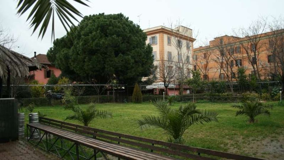 Giardino urbano con prato e piccole palme, una lunga panchina in legno in primo piano e un grande albero sempreverde al centro. Sullo sfondo si vedono edifici residenziali di più piani e alberi spogli lungo il perimetro.