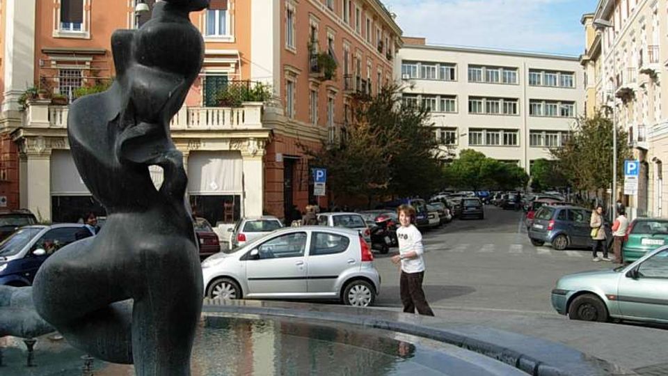 Fontana circolare in una piazza urbana, con una grande statua scura al centro e vasca d’acqua in primo piano. Sullo sfondo si vedono palazzi multipiano con facciate color pastello, strada con auto parcheggiate e alcuni alberi lungo i marciapiedi.