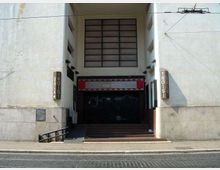 Ingresso incassato in un edificio chiaro con grande finestra rettangolare a griglia sopra e portone scuro chiuso, raggiunto da una breve scalinata e una rampa laterale. Ai lati dell’entrata sono presenti insegne verticali e pareti in muratura con segni di usura.