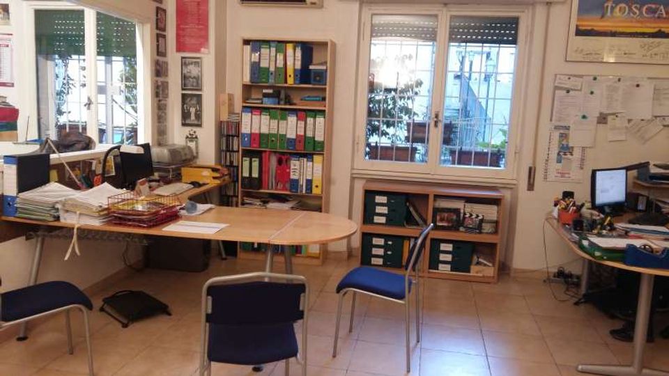 Interno di un ufficio con scrivanie disposte ai lati, sedie blu al centro e scaffali con numerosi raccoglitori colorati. Due grandi finestre con tapparelle parzialmente abbassate illuminano la stanza; sulle pareti ci sono bacheche e poster, con computer e documenti su una postazione laterale.