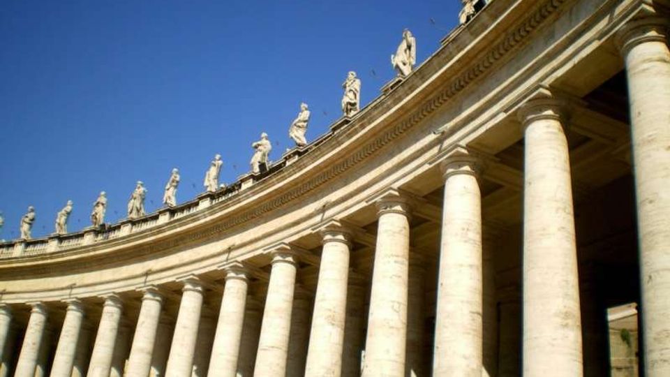 Colonnato monumentale in pietra con fila di alte colonne e trabeazione curva, sormontato da statue lungo la sommità. Sullo sfondo si vede un cielo azzurro limpido.