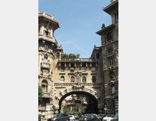 Veduta stradale urbana con palazzi storici in pietra riccamente decorati e un grande arco monumentale che collega due edifici sopra la carreggiata. Si notano balconi, finestre con cornici ornate e sculture sulla facciata, con automobili parcheggiate e cielo sereno sullo sfondo.