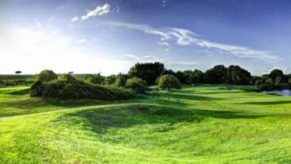 Ampio paesaggio panoramico di un campo da golf con prati ondulati e alberi sparsi, sotto un cielo azzurro con nuvole sottili. Sul lato destro si vede uno specchio d’acqua che costeggia il green.