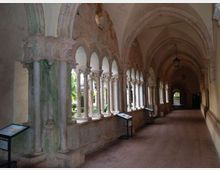 Corridoio di un chiostro storico con volte a crociera e una lunga serie di archi sorretti da colonnine in pietra, alcune con fusto tortile. Sul lato aperto si intravede il giardino interno e lungo il percorso sono presenti pannelli informativi su supporti metallici.