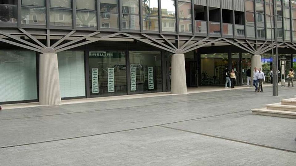 Facciata di un edificio moderno con ampie vetrate a griglia e colonne cilindriche che sorreggono una struttura a travi incrociate. Davanti si apre una grande piazza pavimentata in cemento, con ingresso coperto e vetrine al piano terra.